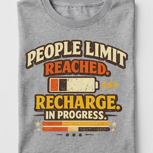 May include: A heather gray t-shirt featuring a graphic with the text "PEOPLE LIMIT REACHED. RECHARGE. IN PROGRESS." The design includes a battery icon with a low charge level, three lightning bolts, two horizontal bars, and three small stars.