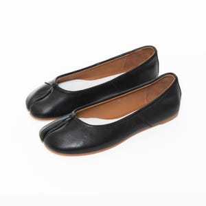 May include: A pair of black leather ballet flats with a split-toe design. The shoes have a small bow detail at the top and a brown sole. The interior is white. The shoes are displayed on a white background.