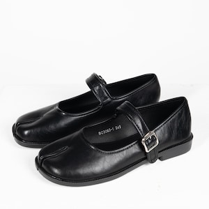Puede incluir: Un par de zapatos Mary Jane de cuero negro con diseño de puntera dividida. Cada zapato tiene una correa con cierre de hebilla y un tacón pequeño. Los zapatos se muestran sobre un fondo blanco.