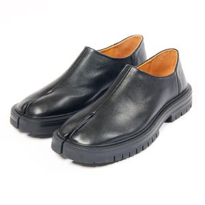 Op de afbeelding: Een paar zwarte leren schoenen met een opvallend ontwerp met gespleten neus, een glad bovenoppervlak en een dikke, zwarte rubberen zool met profiel. De binnenvoering van de schoenen is uitgevoerd in een lichtbruine kleur.