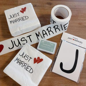 Könnte beinhalten: Hochzeitsdekorationen mit dem Schriftzug "Just Married" in schwarzer Schrift und roten Herzakzenten. Zu den Artikeln gehören Servietten, Bänder und ein Banner. Eine Rolle weißes Klebeband mit dem gleichen Text ist ebenfalls sichtbar.