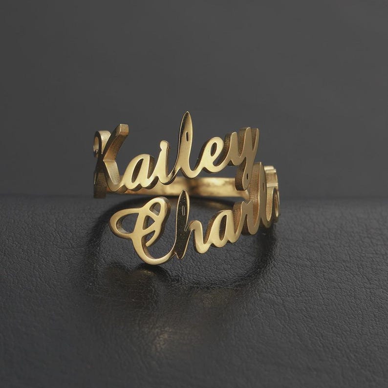 May include: A gold-colored ring with the names "Kailey" and "Charlie" in a cursive font. The ring is displayed on a dark surface, with the names overlapping. The ring is a personalized jewelry item.