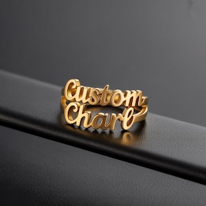 May include: A gold-colored ring with the words "Custom" and "Charle" in a cursive font. The ring has a double-band design, with the names stacked on top of each other. The ring is displayed on a black surface.