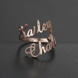 May include: A rose gold ring with the names "Karley" and "Charli" in a cursive font. The ring is displayed on a dark surface, showcasing its elegant design and personalized touch. A unique piece of jewelry.