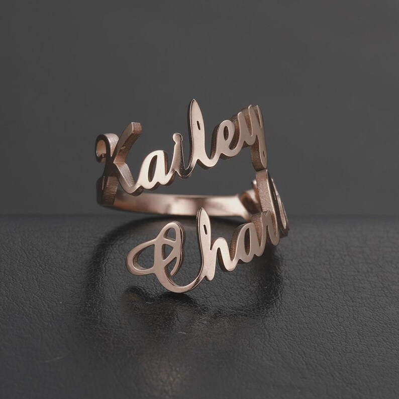 May include: A rose gold ring with the names "Karley" and "Charli" in a cursive font. The ring is displayed on a dark surface, showcasing its elegant design and personalized touch. A unique piece of jewelry.