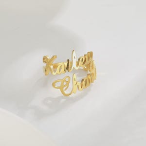 May include: A gold-colored ring with the names "Kailey" and "Chana" in a cursive font. The ring is open and wraps around the finger. The ring is on a white surface.