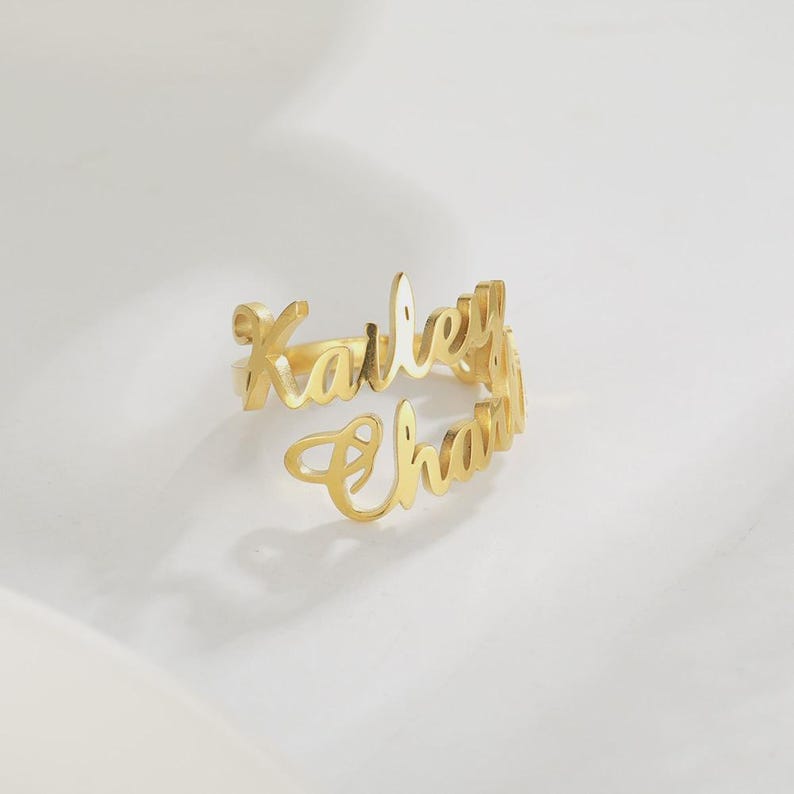May include: A gold-colored ring with the names "Kailey" and "Chana" in a cursive font. The ring is open and wraps around the finger. The ring is on a white surface.