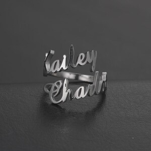 May include: A silver-colored ring with the names "Kailey" and "Charlie" in a cursive font. The ring has a double-band design, with each name forming a separate band that wraps around the finger. The ring is displayed on a dark surface.