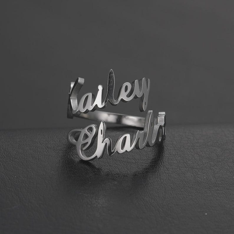 May include: A silver-colored ring with the names "Kailey" and "Charlie" in a cursive font. The ring has a double-band design, with each name forming a separate band that wraps around the finger. The ring is displayed on a dark surface.