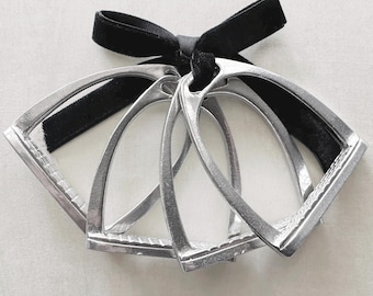 May include: Three silver-toned metal stirrups arranged in a fan-like formation, each featuring a textured tread plate at the base. The stirrups are held together at the top by a black velvet ribbon tied into a simple bow, creating a cohesive set.