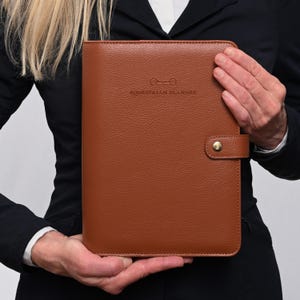 May include: A tan, textured leather-like planner with a gold-tone snap closure. The front cover features a debossed logo of a bit and the text EQUESTRIAN PLANNER. The rectangular organizer has rounded corners and a professional, structured appearance.