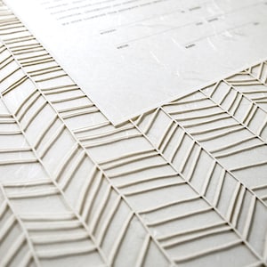 May include: A close-up of a white paper with a chevron pattern embossed on the surface. The paper is textured and has a subtle sheen.