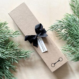 May include: A brown gift box with a black ribbon and bow. The box has a white label with the text "Jennifer Baichman" and a small black and white illustration of a horse bit.