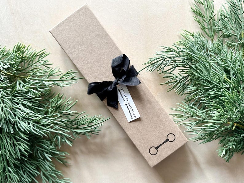 May include: A brown gift box with a black ribbon and bow. The box has a white label with the text "Jennifer Baichman" and a small black and white illustration of a horse bit.