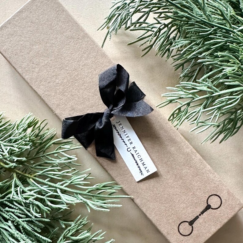 May include: A brown paper gift box with a black ribbon and a gift tag that reads "Jennifer Raichman". The box is surrounded by green foliage.