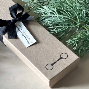 May include: A brown cardboard box with a black ribbon tied around it. The box has a white label with the text "Jennifer Raichman" and a small horse bit symbol.