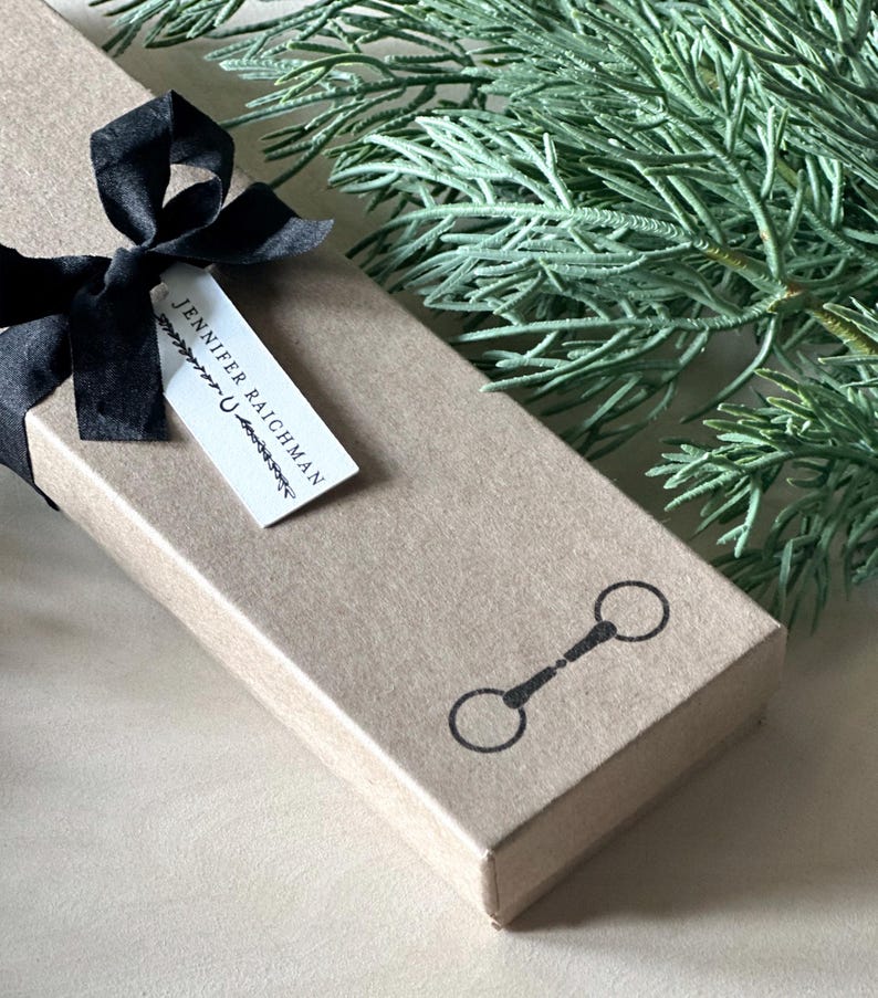 May include: A brown cardboard box with a black ribbon tied around it. The box has a white label with the text "Jennifer Raichman" and a small horse bit symbol.