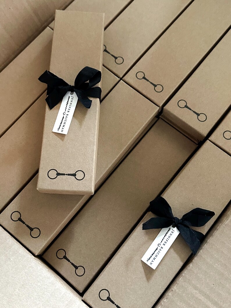 May include: A stack of brown cardboard boxes with black ribbon bows and a small white tag with the text "Jennifer Rauchman" on each box. The boxes have a black horseshoe design printed on the front.