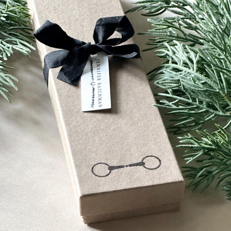May include: A brown cardboard gift box with a black ribbon tied around it. The box has a black and white horse bit design on the front and a white gift tag with the text "Jennifer Raichman" on it.