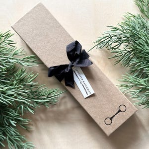 May include: A brown gift box with a black ribbon and a white tag that says "Jennifer Raichman". The box is surrounded by green foliage.
