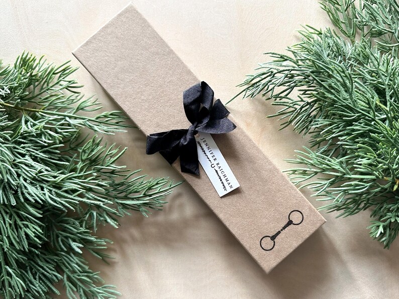 May include: A brown gift box with a black ribbon and a white tag that says "Jennifer Raichman". The box is surrounded by green foliage.