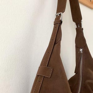 May include: A brown suede shoulder bag with a silver zipper and buckle closure. The bag has a long, adjustable strap and is hanging against a white wall. The bag is a triangular shape.