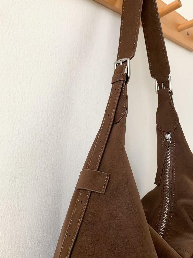 May include: A brown suede shoulder bag with a silver zipper and buckle closure. The bag has a long, adjustable strap and is hanging against a white wall. The bag is a triangular shape.