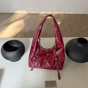 Puede incluir: Un bolso de hombro de cuero burdeos con asa curva y correas ajustables. El bolso tiene hebillas plateadas y se exhibe sobre una superficie blanca. Dos jarrones negros y redondeados est&aacute;n a cada lado del bolso.