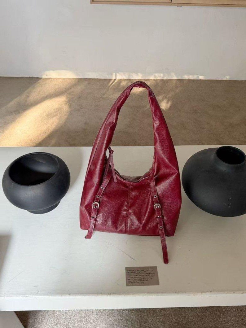 Puede incluir: Un bolso de hombro de cuero burdeos con asa curva y correas ajustables. El bolso tiene hebillas plateadas y se exhibe sobre una superficie blanca. Dos jarrones negros y redondeados est&aacute;n a cada lado del bolso.