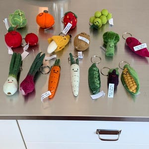 May include: A collection of felt food keychains, including a carrot, onion, banana, strawberry, grapes, corn, potato, broccoli, radish, pea pod, and a green and red cabbage. The keychains are all handmade and feature cute faces.