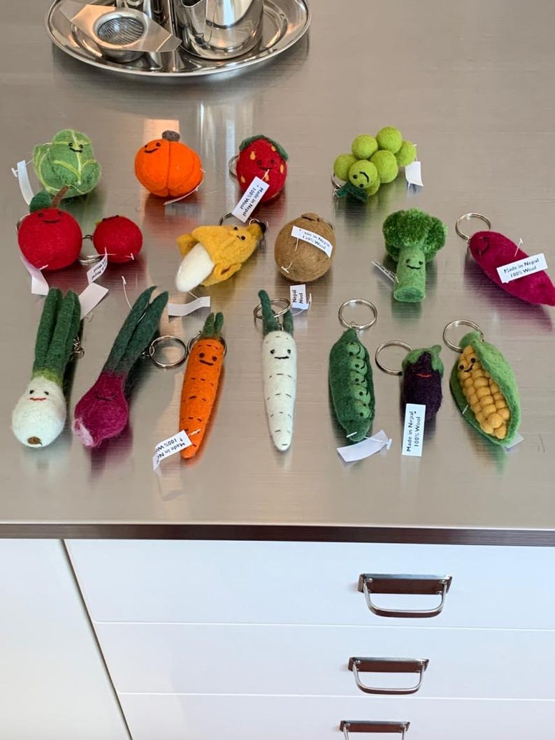 May include: A collection of felt food keychains, including a carrot, onion, banana, strawberry, grapes, corn, potato, broccoli, radish, pea pod, and a green and red cabbage. The keychains are all handmade and feature cute faces.