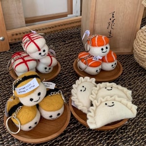 May include: A collection of handmade felt keychains shaped like sushi, dumplings, and other food items. The keychains are white, orange, yellow, and black, with smiling faces. Each keychain sits on a small wooden plate.