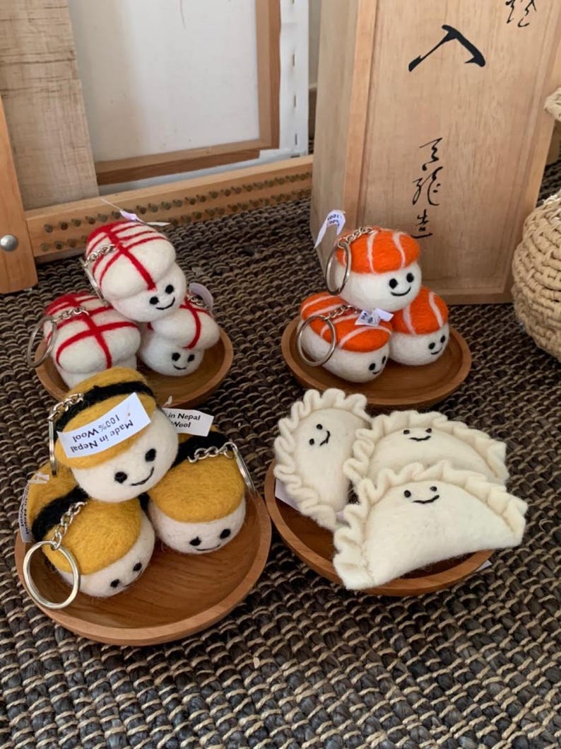 May include: A selection of handmade felt keyrings in the shape of sushi, dumplings, and other food items. The keyrings are white, orange, yellow, and black, with smiling faces. Each keyring sits on a small wooden plate.