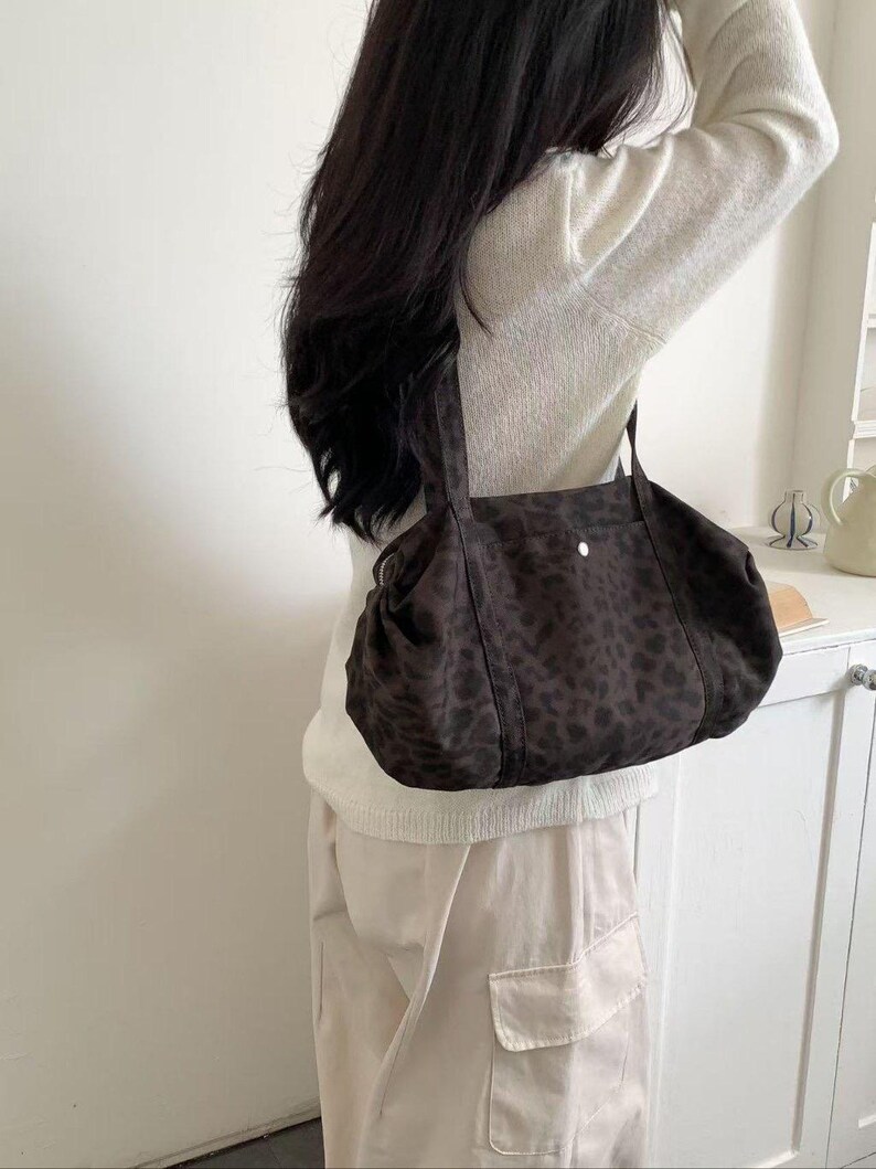 Puede incluir: Un bolso tote con estampado de leopardo marr&oacute;n y negro con dos asas. El bolso est&aacute; hecho de un material suave y tiene un cierre de bot&oacute;n. El bolso se lleva al hombro.