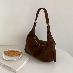 May include: A brown suede shoulder bag with a curved design and a silver zipper. The bag has a long, adjustable strap and a small loop detail. A croissant sits on a white plate next to a book.