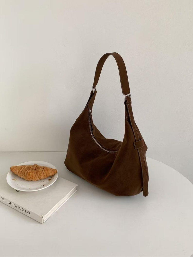 May include: A brown suede shoulder bag with a curved design and a silver zipper. The bag has a long, adjustable strap and a small loop detail. A croissant sits on a white plate next to a book.