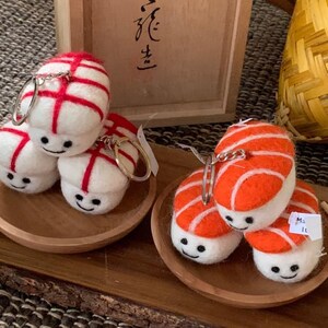 May include: Sushi-shaped keyrings. The keyrings are white with red or orange detailing, and each has a smiling face. They are presented on small wooden plates.