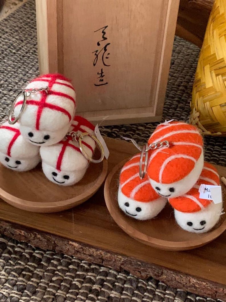 May include: Sushi-shaped keyrings. The keyrings are white with red or orange detailing, and each has a smiling face. They are presented on small wooden plates.