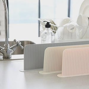 K&ouml;nnte beinhalten: Drei rechteckige Sp&uuml;lbecken-Spritzschutzvorrichtungen aus Silikon in Grau, Beige und Hellrosa. Jeder Schutz verf&uuml;gt &uuml;ber eine vertikal gerippte Textur und eine flache Basis f&uuml;r die Arbeitsplatte. Diese flexiblen Barrieren stehen in einer Reihe am Wasserhahn.
