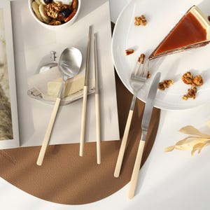 Puede incluir: Un juego de cubiertos de plata con mangos de color crema. El juego incluye una cuchara, un tenedor y palillos. Los cubiertos están dispuestos sobre una superficie de cuero marrón.