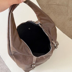 Puede incluir: Un bolso hobo de cuero marr&oacute;n con un asa curva y cierre de cremallera. El bolso tiene un interior negro y herrajes plateados. El bolso tiene un detalle de hebilla en el lateral.