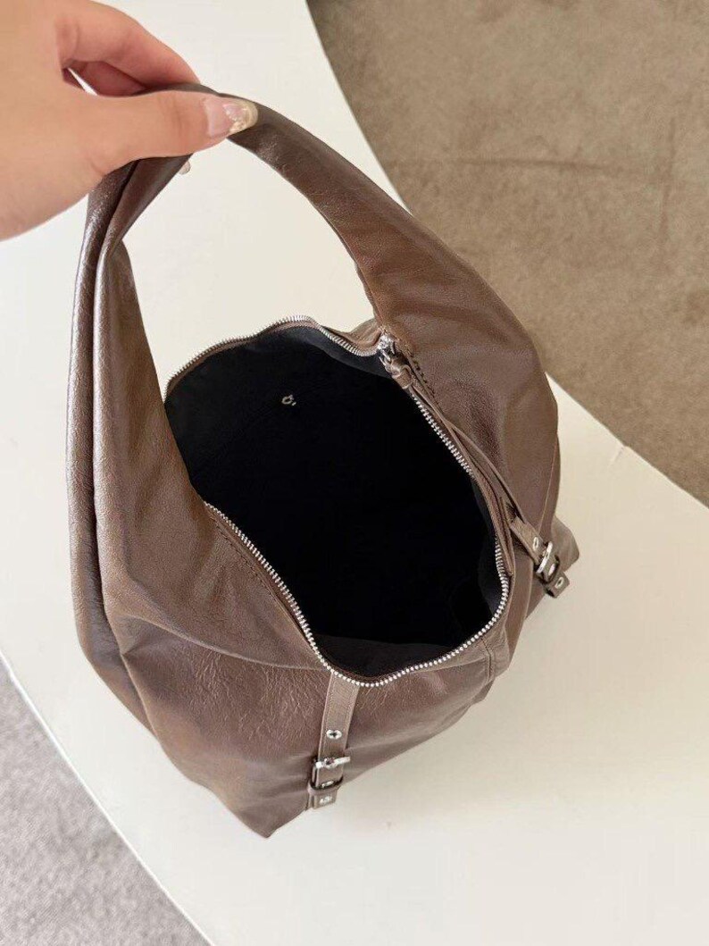 Puede incluir: Un bolso hobo de cuero marr&oacute;n con un asa curva y cierre de cremallera. El bolso tiene un interior negro y herrajes plateados. El bolso tiene un detalle de hebilla en el lateral.