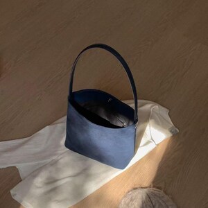 May include: A navy blue handbag with a single handle sits on a white garment. The bag has a rounded shape and a dark interior. A light-colored knit beanie is in the foreground. The items are on a light-colored wooden surface.