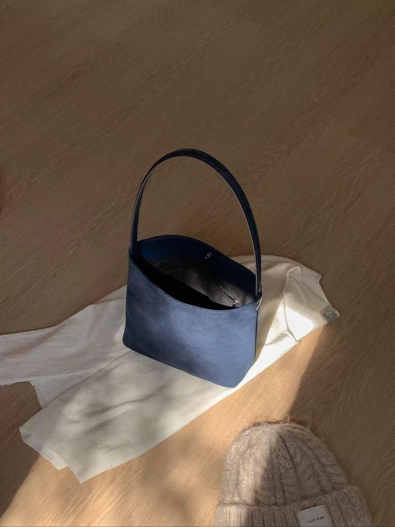 May include: A navy blue handbag with a single handle sits on a white garment. The bag has a rounded shape and a dark interior. A light-colored knit beanie is in the foreground. The items are on a light-colored wooden surface.