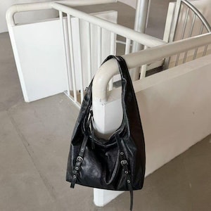Puede incluir: Un bolso de hombro de cuero negro con un dise&ntilde;o holgado. El bolso tiene correas ajustables con hebillas plateadas y una sola correa para el hombro. El bolso est&aacute; colgado de una barandilla blanca en un espacio de tonos neutros.