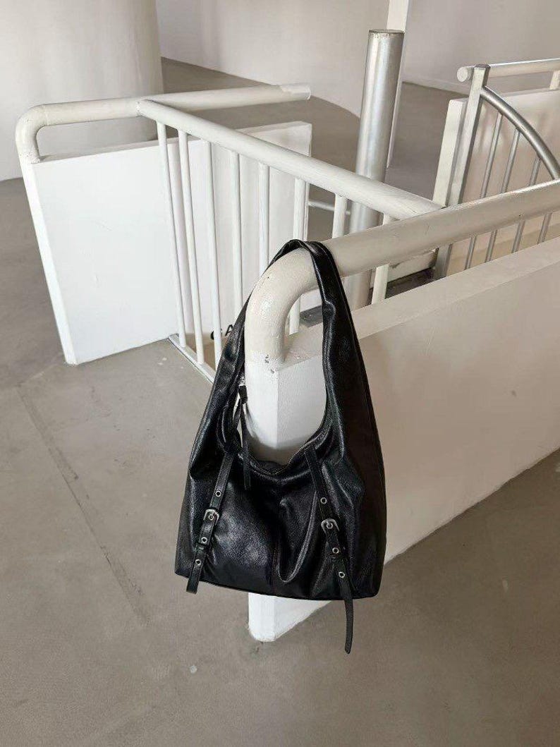 Puede incluir: Un bolso de hombro de cuero negro con un dise&ntilde;o holgado. El bolso tiene correas ajustables con hebillas plateadas y una sola correa para el hombro. El bolso est&aacute; colgado de una barandilla blanca en un espacio de tonos neutros.