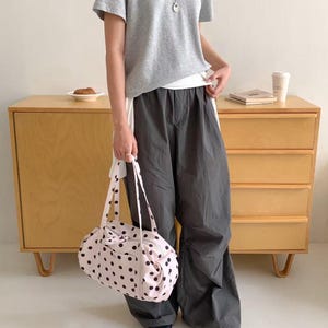 Puede incluir: Una camiseta gris claro y pantalones grises oscuros se combinan con un bolso de mano rosa claro con lunares. El bolso tiene lunares negros y correas largas. Tambi&eacute;n se ve un collar plateado.