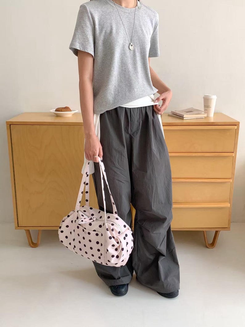 Puede incluir: Una camiseta gris claro y pantalones grises oscuros se combinan con un bolso de mano rosa claro con lunares. El bolso tiene lunares negros y correas largas. Tambi&eacute;n se ve un collar plateado.