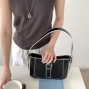 May include: Black handbag with white edging and a silver buckle. The bag is rectangular with a handle. A silver necklace with a pendant is also visible. A cup and saucer are on the table.