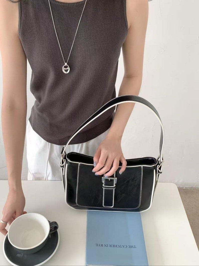 May include: Black handbag with white edging and a silver buckle. The bag is rectangular with a handle. A silver necklace with a pendant is also visible. A cup and saucer are on the table.